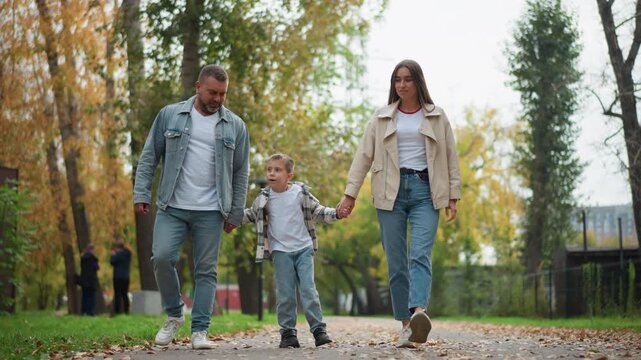 Family of three walking together in park holding hands and enjoying time outdoors. man wears denim jacket over white shirt with jeans woman has on beige coat above her own t shirt paired