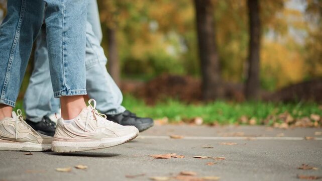 Close up of feet in motion wearing casual sneakers and jeans on paved path with fallen leaves. Multiple pairs visible showing different shoe styles including white mesh athletic shoes worn