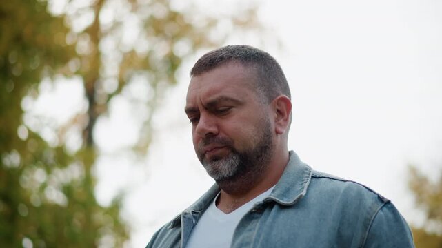 Close up of man wearing denim jacket and white t shirt standing outdoors with trees in background. Man maintains neutral expression throughout head slightly tilted downward. Soft natural