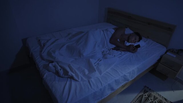 Man doomscrolling on smartphone in bed late at night, delaying sleep due to phone addiction and bedtime procrastination