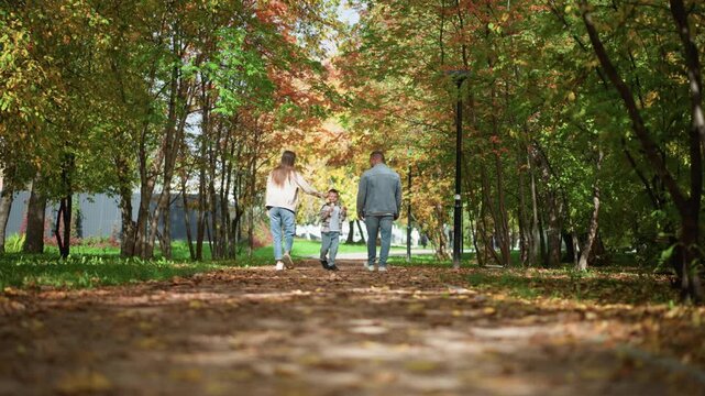 Family of three walking together along leaf strewn path in autumn park with colorful foliage and soft natural light. Child holds hands between adult pair all wearing casual clothing