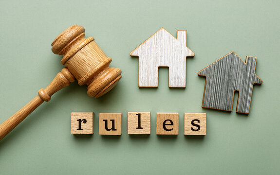 Wooden judge gavel next to letter blocks spelling rules and small house models on green background symbolizing real estate law and property code