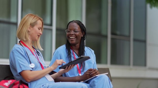 Two female nurses discussing medical records on a tablet outside the hospital