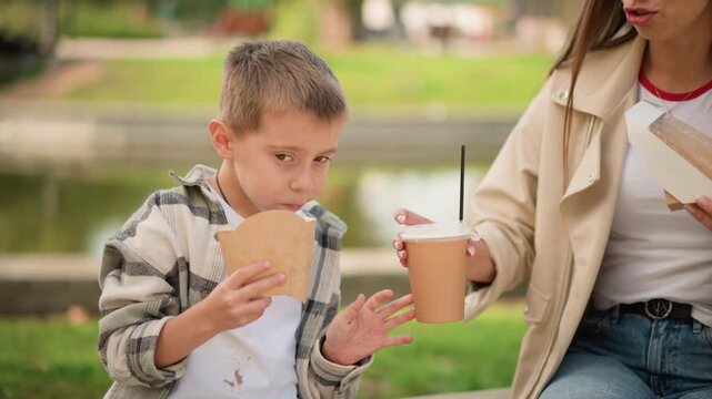 Close up of young boy enjoying ice cream while sitting outdoors with woman. Boy holds paper cup and wrapper interacting naturally as they share moment in park setting near water feature