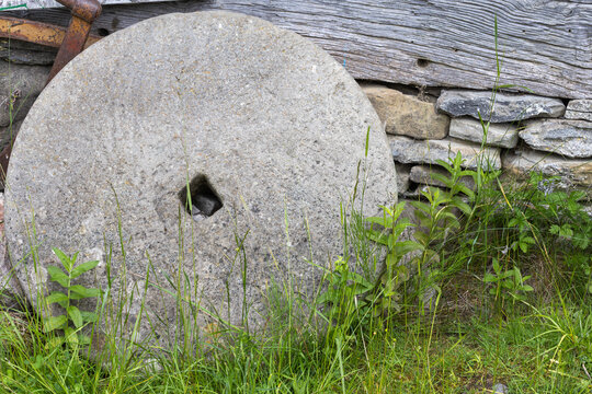 a historic circular grindstone resting in green overgrowth, positioned against a rural exterior wall constructed of stacked rock and aged timber.