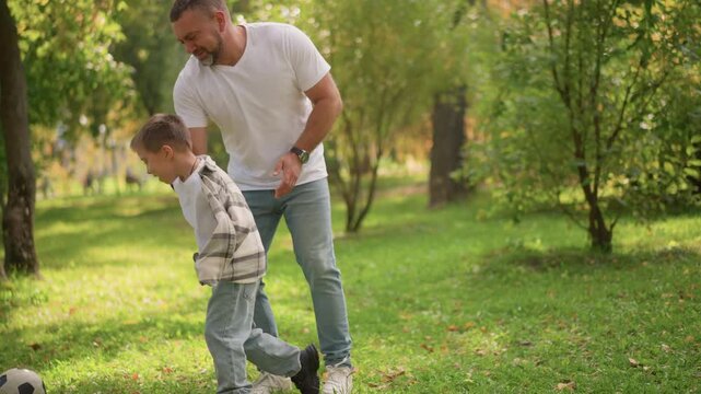Young boy in plaid jacket plays soccer with adult man on grassy ground. child dribbles ball while another boy joins them all set against green park surroundings with trees and scattered
