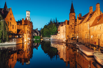 Obraz premium Historic medieval city of Bruges warmly illuminated against blue sky, mirrored in the calm waters of the canal.