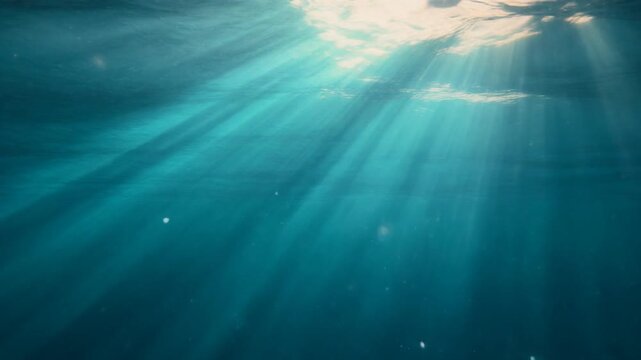 Sunlight rays penetrate the turquoise underwater surface viewed from below, illuminating tiny suspended particles in the.