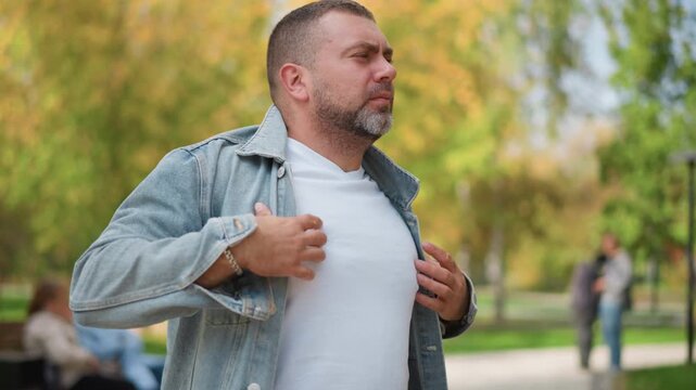 Close up of man putting on denim jacket over white t shirt in park setting with blurred people and greenery background. Man adjusts clothing while standing outdoors showing casual attire