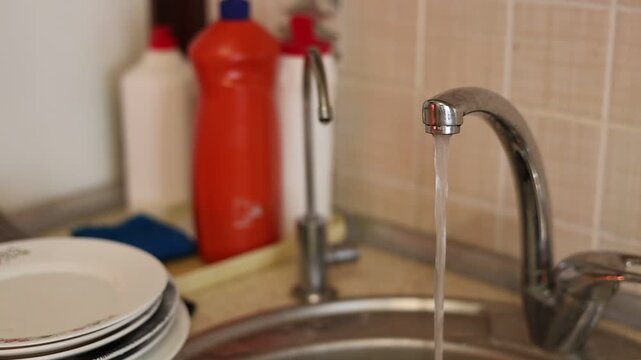 Running water from a kitchen faucet filling a bowl in a stainless steel sink with dirty dishes piled on the countertop, household chores and cleaning concept