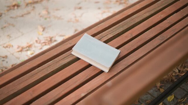 Close up of book resting on wooden slats with fallen leaves in background. scene is calm and still focusing on texture detail and soft lighting. single white covered object sits stationary