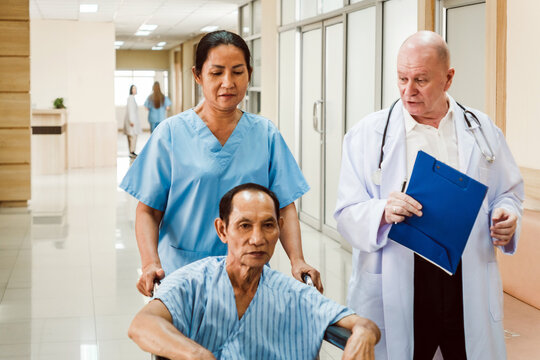 Doctor with clipboard speaks to a nurse pushing a patient in a wheelchair down a bright hospital corridor
