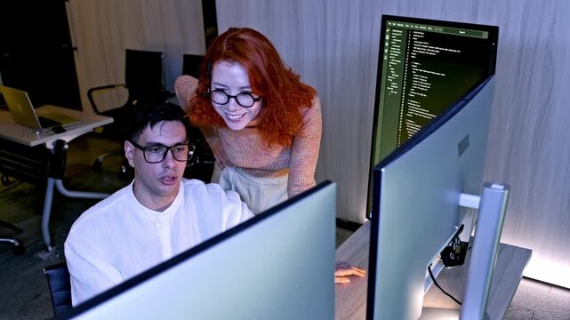 Two developers collaborate at dual monitors in a modern office, reviewing code and discussing solutions.