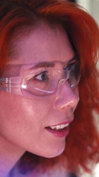 Close-up profile of a person with red hair wearing clear safety glasses, focused under lab lighting.