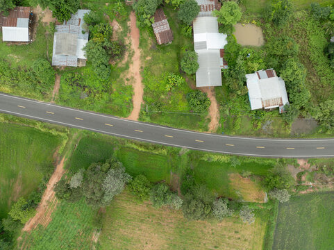 Aerial view of rural asphalt road passing green rice fields and farmhouses during rainy season. Rural infrastructure that connects farming communities, agriculture and local food production.