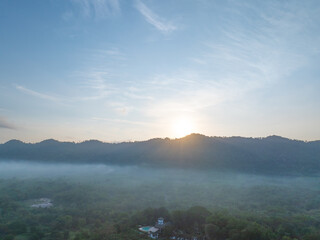 Sunrise emerging behind a forested mountain ridge above misty tropical countryside, with golden morning light illuminating low fog and lush woodland for a peaceful nature. Travel and wellness themes. © Fahroni