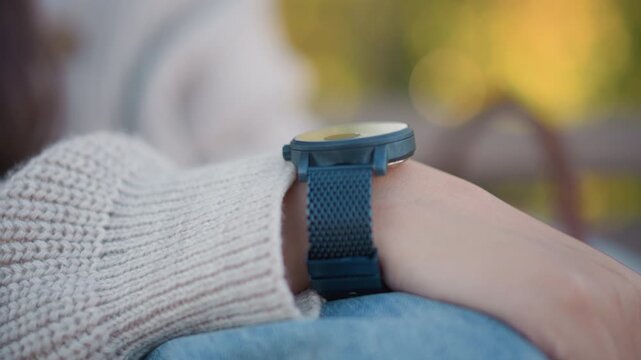 Close up of woman wrist wearing blue watch with fabric strap resting on lap. Knitted sweater sleeve and soft focus background with warm tones visible knit