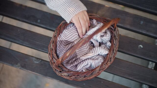Close up of wicker basket filled with folded chevron patterned blankets on wooden slats. person hand in gray knitwear reaches towards blanket then rests inside basket. scene is softly lit