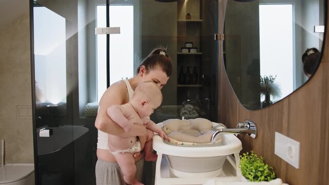 Woman holding infant near baby bathtub and sink in contemporary bathroom setting with wood accents and glass shower