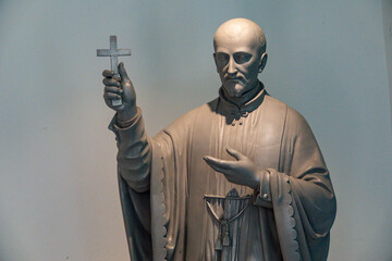A statue of Saint John Francis Regis stands indoors holding a small raised metal crucifix. © Joan