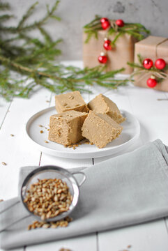 Sunflower halva pieces on a white plate with seeds, fir branches, red ornaments, and Christmas gifts in the background.