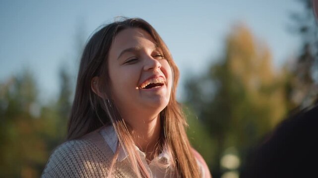 Close up of young woman with long hair smiling outdoors in natural light. Woman wears knitted sweater over collared shirt interacting naturally against outdoor backdrop with trees and sky