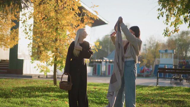 Two women interact in park setting one holding wicker basket while other spreads out patterned blanket. scene features autumn foliage with golden leaves and green grass under bright sunlight