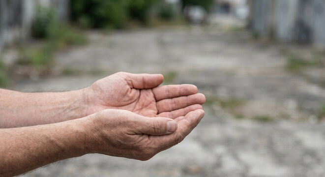Hands of a middle-aged man with weathered skin extended outward, palms up, against a blurred urban background with concrete pavement and greenery