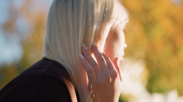 Close up of young woman with long blonde hair adjusting her face in soft sunlight. Woman wears dark clothing and interacts naturally moving slightly. background features blurred autumn