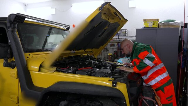 Mechanic checks engine under yellow off-road vehicle. Workshop lights shine brightly on tools and equipment. Technician wears high-visibility uniform, focused on task. Ideal for automotive repair