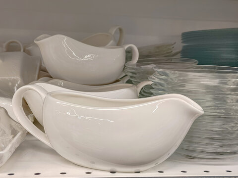 Ceramic gravy boat on store shelf with stacks of glass plates. Tableware retail display in kitchenware department. Household goods shopping for dining room and home kitchen equipment