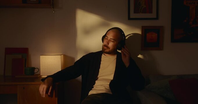 A bald bearded man wearing headphones relaxes with closed eyes in a cozy room illuminated by a warm table lamp light and dramatic golden evening sunlight, enjoying music in peaceful solitude.