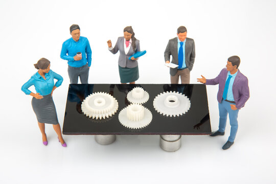 Miniature people discuss gears on a table in a bright room