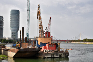 Fototapeta premium Construction Barges and Heavy Cranes on Riverfront Near Skyscraper