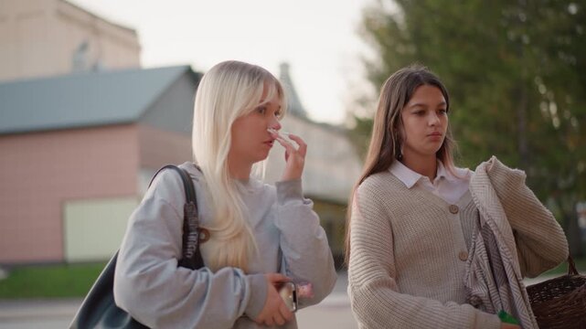 Two young women walk together outdoors one holding phone and other carrying woven basket. They wear casual clothing including sweaters with visible textures like knitted patterns on their
