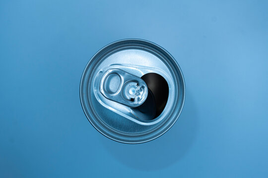 Top view of an opened aluminum beverage can against a clean blue background. The metallic pull tab, circular rim, and dark opening create a simple modern composition with strong visual contrast. Suita