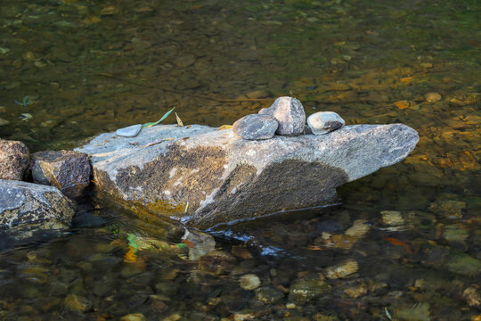 Several smooth river stones are neatly stacked in a small tower on top of a large boulder. The composition is set in shallow, transparent water, allowing pebbles on the bottom to be seen.
