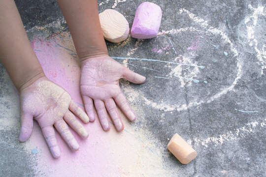 Child's hands covered in colorful chalk dust while drawing on asphalt. Nearby are pieces of sidewalk chalk and the colorful marks they have left behind.