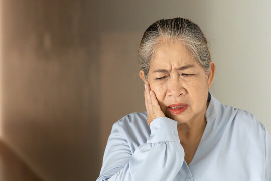 Asian senior, old aged woman suffering from toothache and tooth decay indoors, holding her cheek in pain, dental care concept