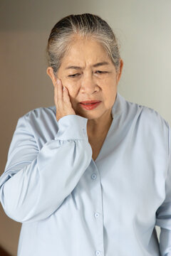 Asian senior, old aged woman suffering from toothache and tooth decay indoors, holding her cheek in pain, dental care concept