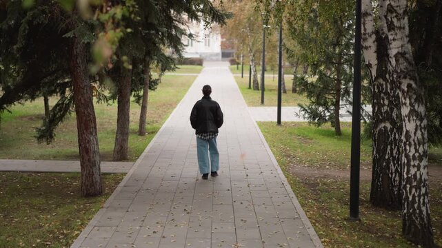 Woman walks steadily down tree lined path in outdoor park wearing casual clothing. scene features fallen leaves on ground and distant buildings visible through trees under soft natural light
