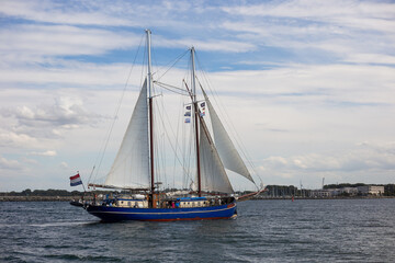 Fototapeta premium Dark blue wooden two-masted schooner with full white sails navigating choppy sea waters during Hanse Sail regatta under fluffy clouds.