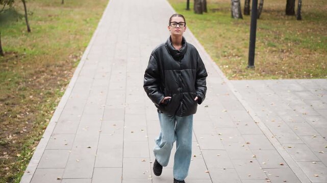 Close up of young woman walking steadily along paved path in outdoor park setting wearing casual attire. Woman maintains hands in pockets posture throughout sequence against grassy areas