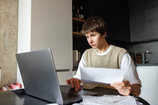 Young focused woman holding financial paper and watching laptop