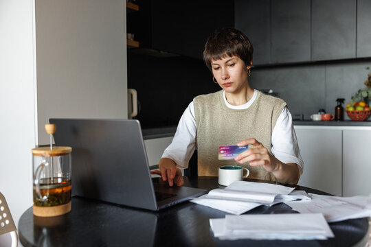Young focused woman paying with credit card on laptop at desk at home