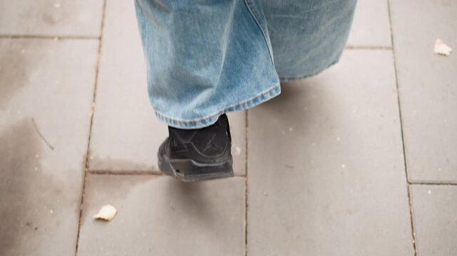Close up of person wearing light blue jeans and black shoes walking on gray tiled pavement with scattered autumn leaves. focus remains steadily on lower legs capturing subtle movement