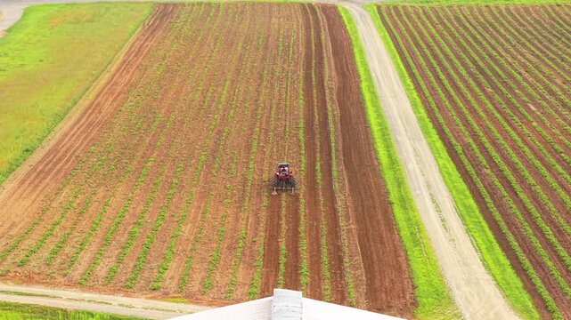 Tractor tilling agricultural crop rows in a green valley field near a rustic barn. Aerial drone tracking video.