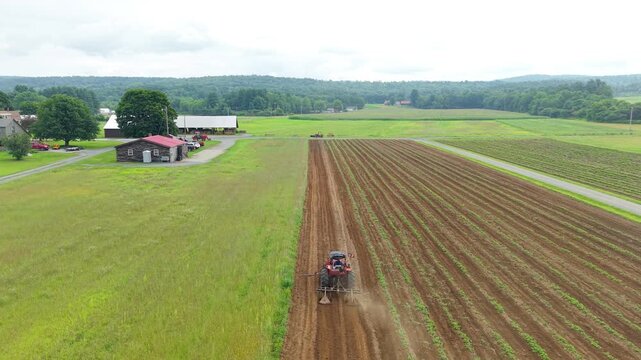 Tractor tilling agricultural crop rows in a green valley field near a rustic barn. Aerial drone tracking video.
