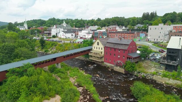 Littleton, New Hampshire, high aerial drone video over the Ammonoosuc River, showing the historic covered bridge, red grist mill, and downtown village architecture set against forested green hills.