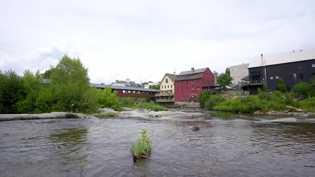 Littleton, New Hampshire, ground-level static shot looking across the flowing Ammonoosuc River toward the historic grist mill, waterwheel, and pedestrian covered bridge under an overcast sky.